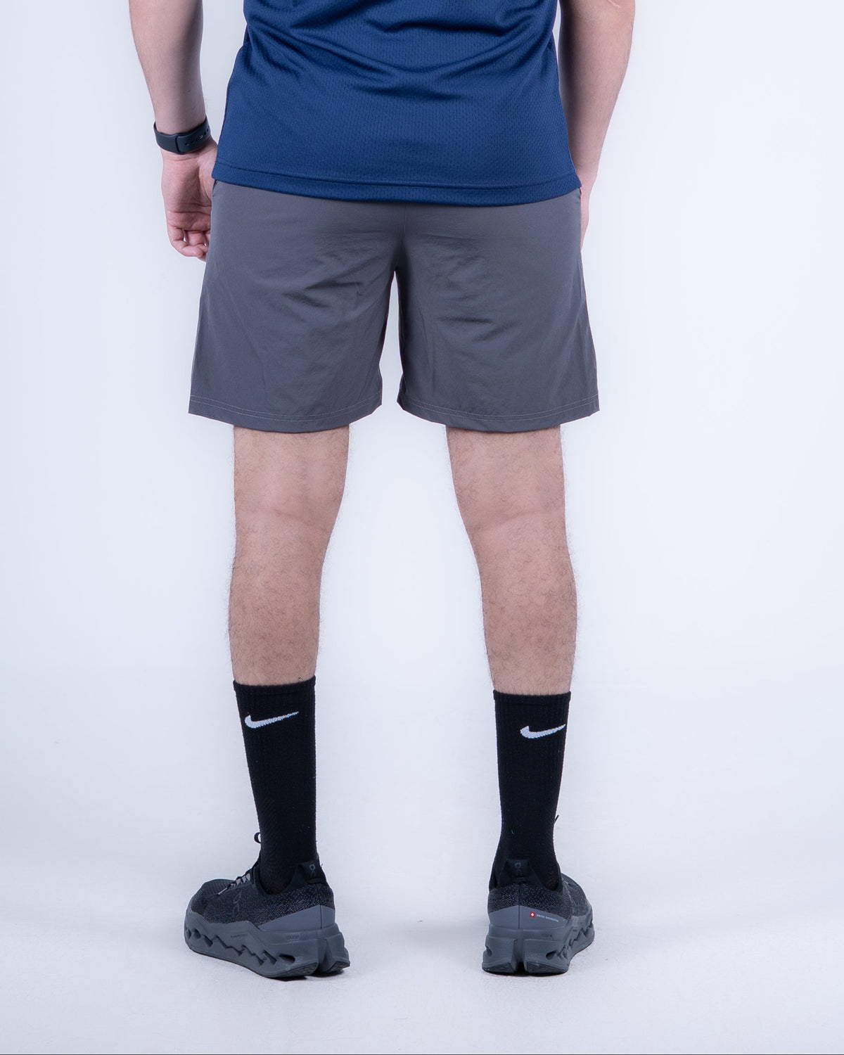 A man is shown from the back wearing a navy blue athletic t-shirt with a small white logo on the upper back, gray running shorts, black Nike socks, and black running shoes, against a light gray studio background.
