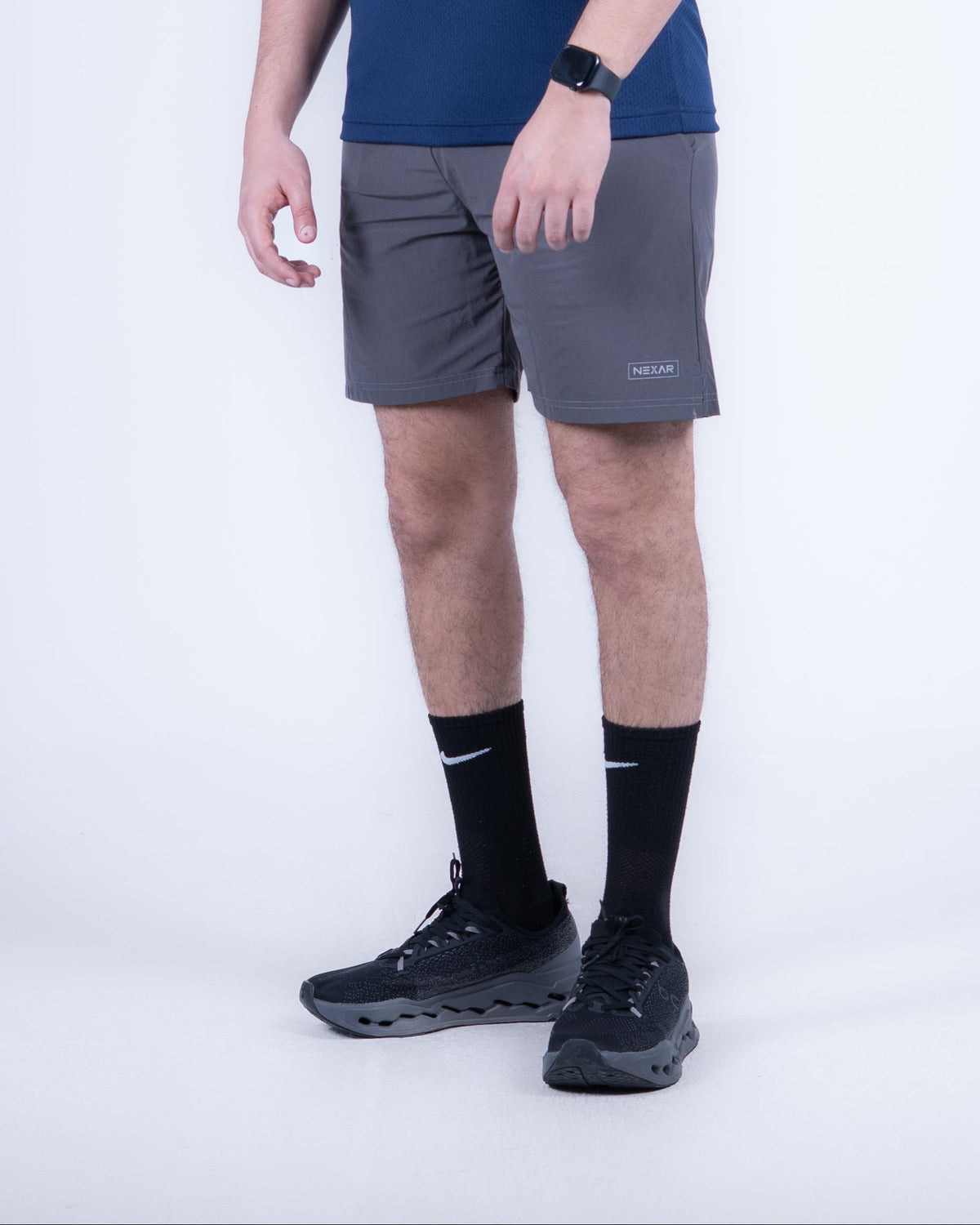 A man modeling a navy blue athletic t-shirt with a small white logo on the left chest, paired with gray athletic shorts, black socks, and black running shoes, against a light gray studio background.