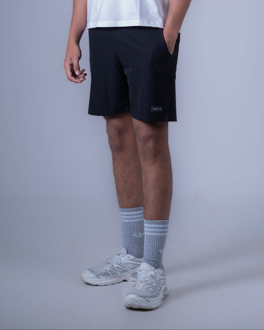 A man modeling a sleeveless white athletic tank top with a small black logo on the chest, paired with black athletic shorts, gray socks, and white sneakers, against a solid gray studio background.